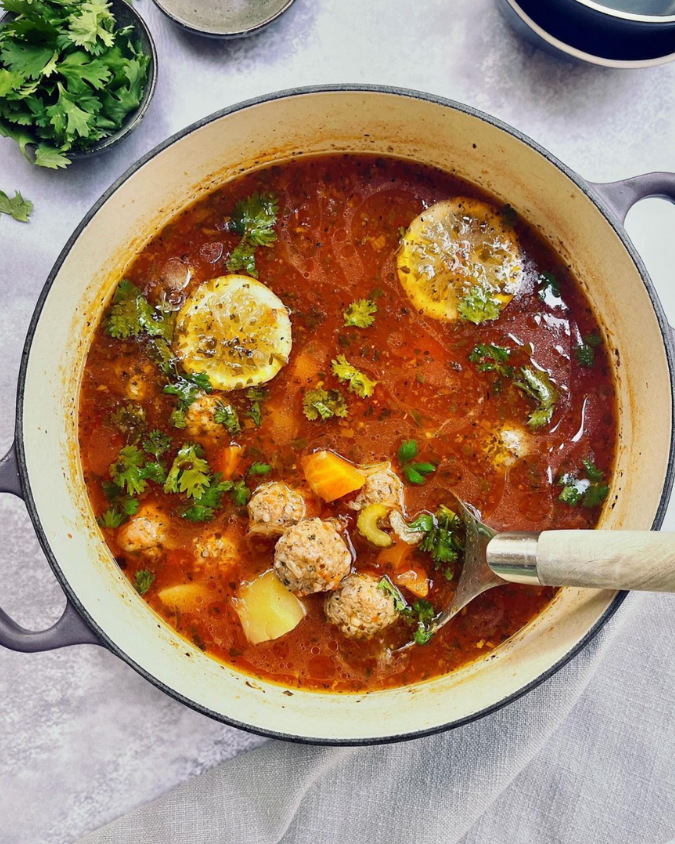 A soup pot filled with rich red colored broth with meatballs, lemon slices, vegetables and fresh herbs.