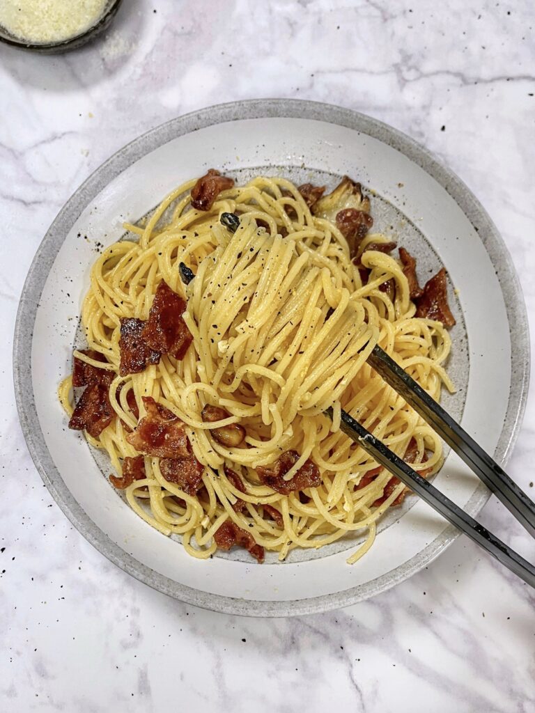 Spaghetti pasta coated in egg and cheese mixture with bacon bits is arranged on a round plate with a light grey background