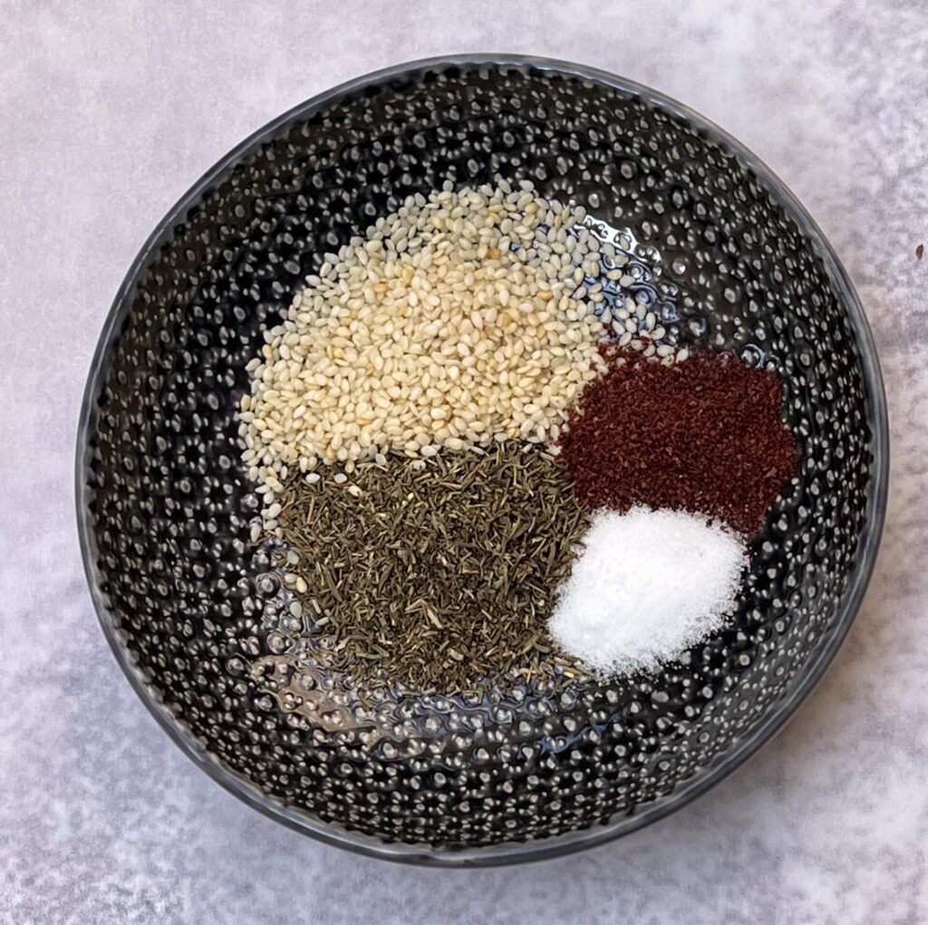 a bowl with ingredients for za'atar seasoning like white sesame seeds, sumac, salt and dried thyme