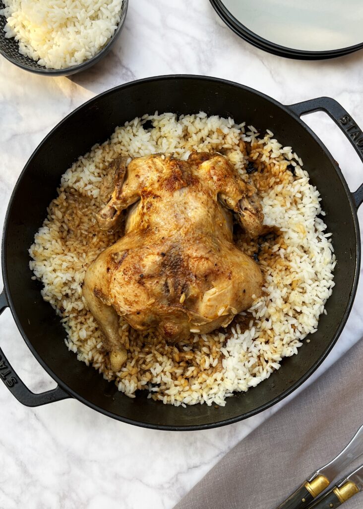 A large casserole with a whole spiced golden brown chicken resting over white rice.