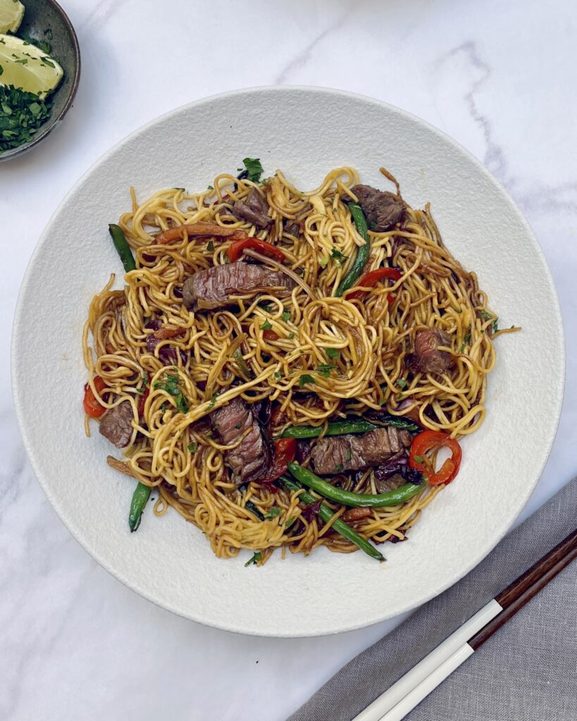 steak noodles in brown sauce with stir fried green and red vegetables and slices of steak cooked to medium