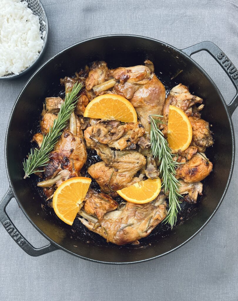 A large casserole with chicken parts coated with a shiny glaze topped with orange slices and rosemary sprigs