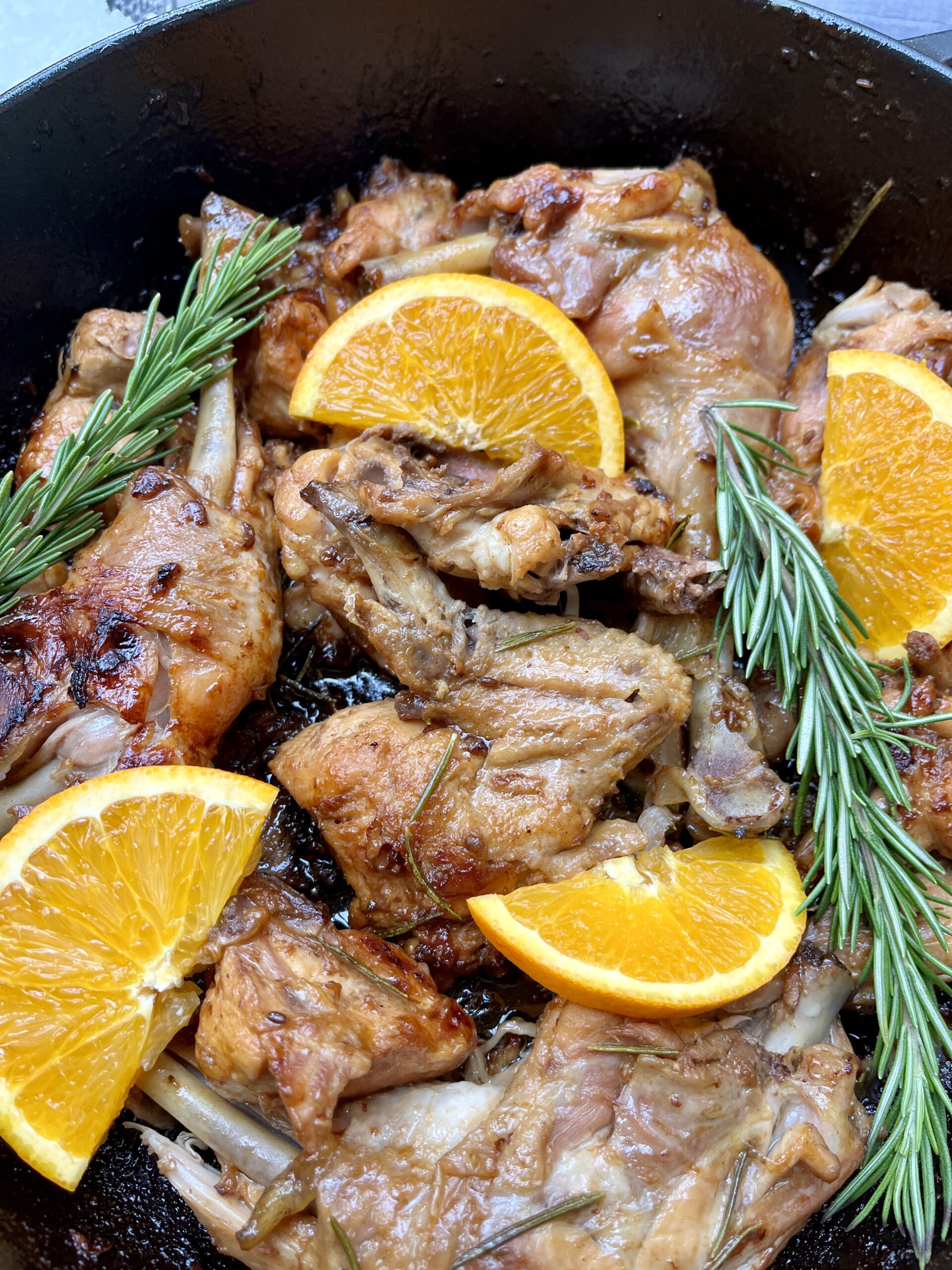 A black casserole with parts of chicken coated in a glaze decorated with orange slices and fresh rosemary