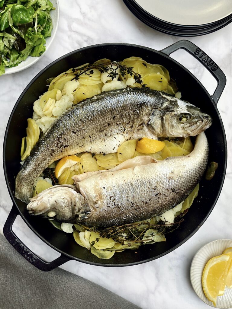 Two whole sea basses lying on a bed of buttery potatoes together with thyme sprigs, lemon slices and garlic cloves