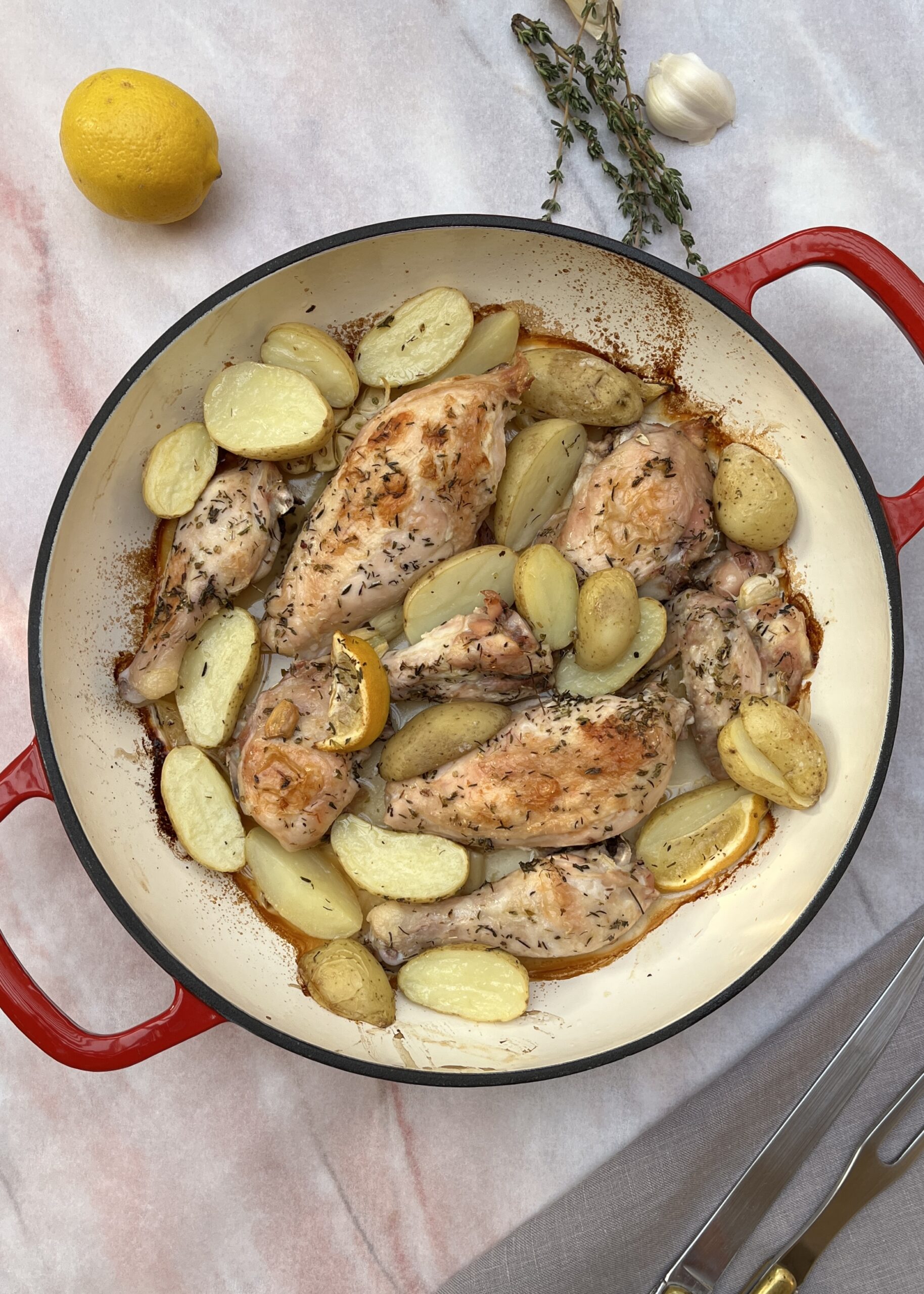 Browned chicken parts in a casserole with halved baby potatoes, lemon wedges, garlic and herbs