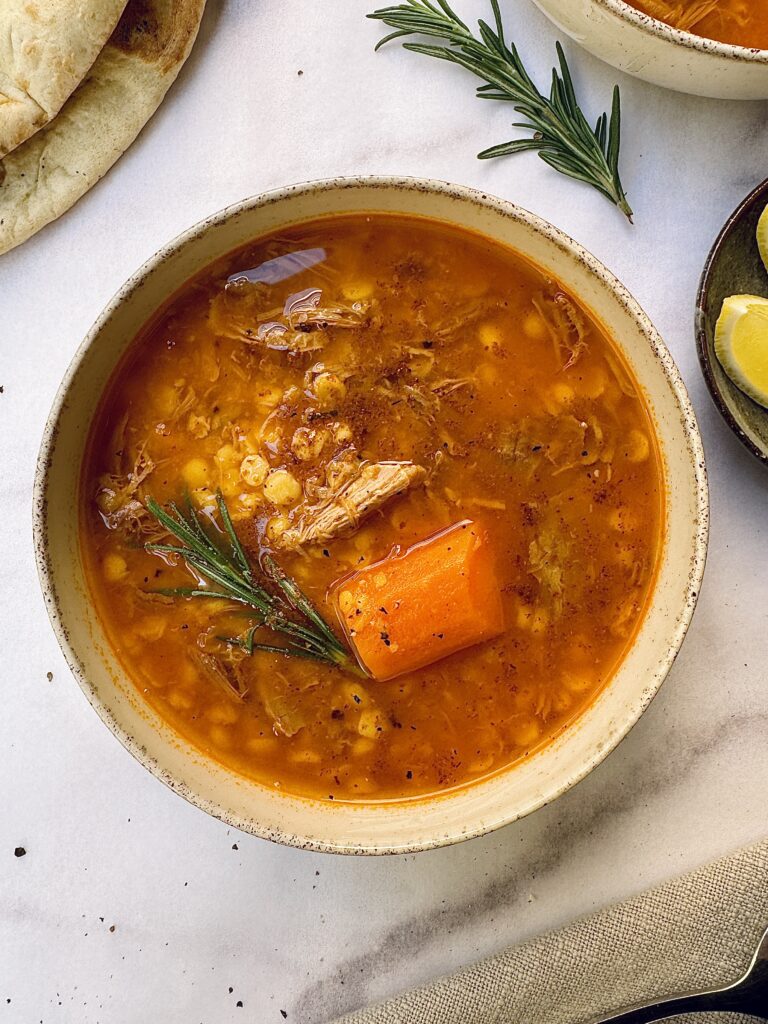 a bowl of soup with a deep red-orange color with chunky carrots, pulled meat and lentils decorated with a rosemary sprig.