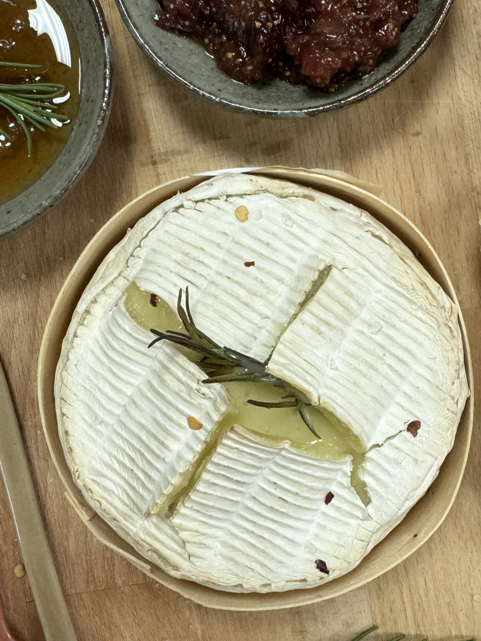 Baked camembert with melting middle decorated with rosemary sprig and crushed chilis sits on a wooden board alongside crusty bread, hobey and fig chutney