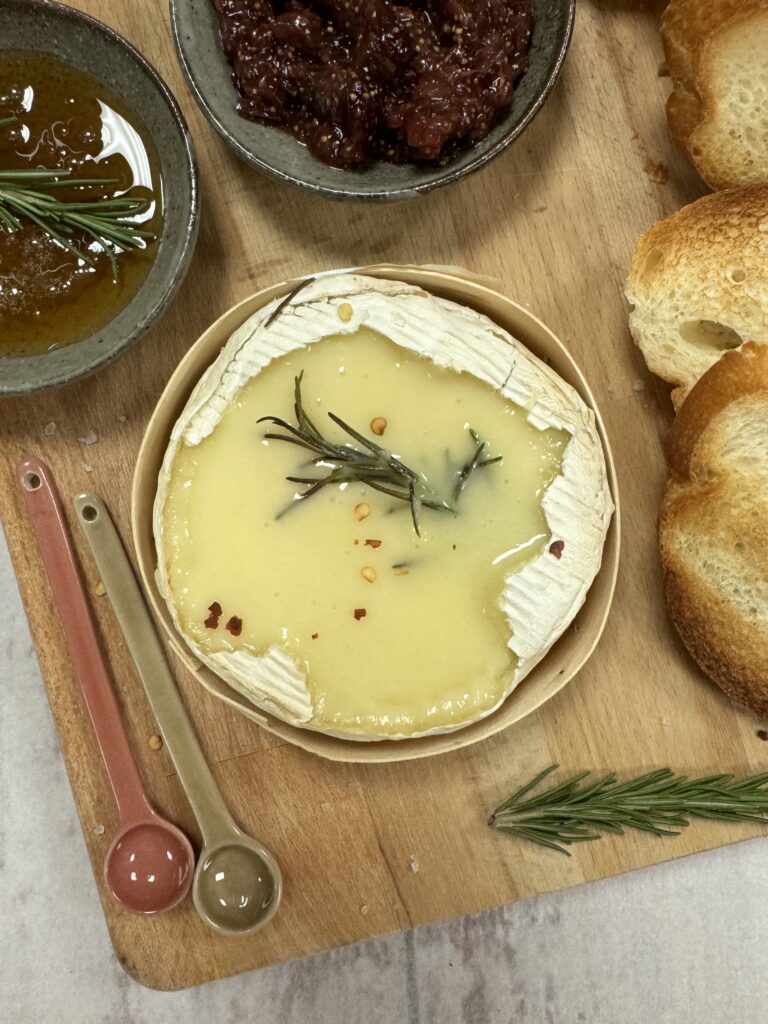 Baked camembert with melting middle decorated with rosemary sprig and crushed chilis sits on a wooden board alongside crusty bread, hobey and fig chutney