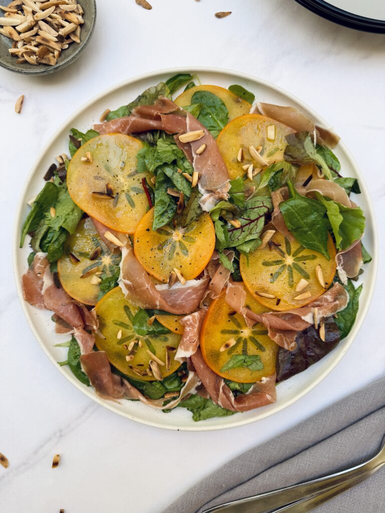 A colorful plate of salad greens topped with sliced persimmon and strips of parma ham topped with toasted almonds.