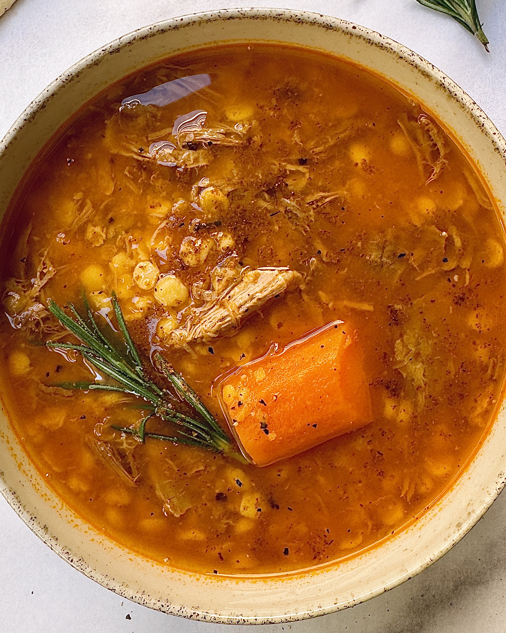 a bowl of soup with a rich red-orange color with a chuncky carrot, pulled meat and lentils decorated with a sprig of rosemary