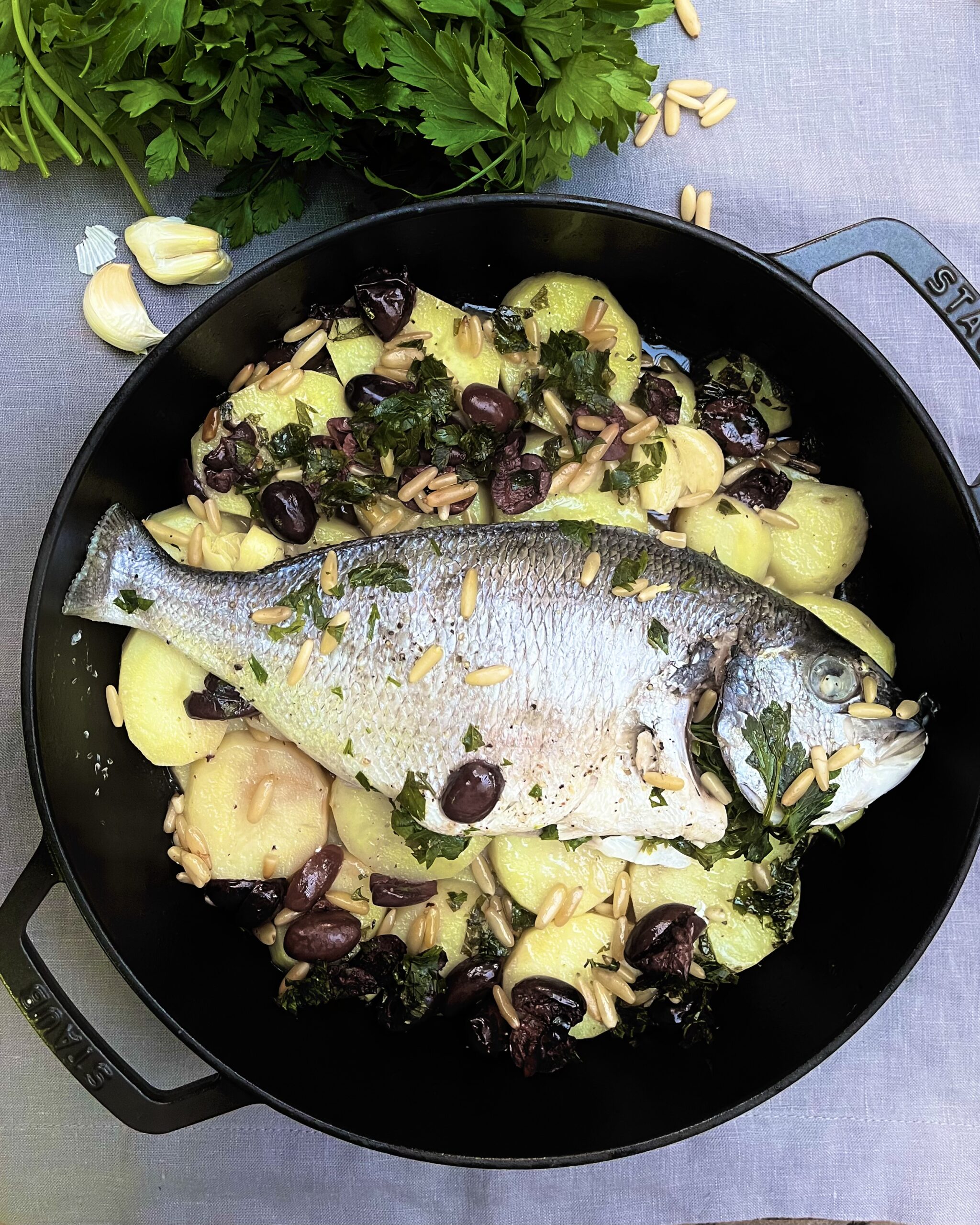 a black casserole with whole sea bream lying on a bed of sliced potatoes with black olives and pine nuts