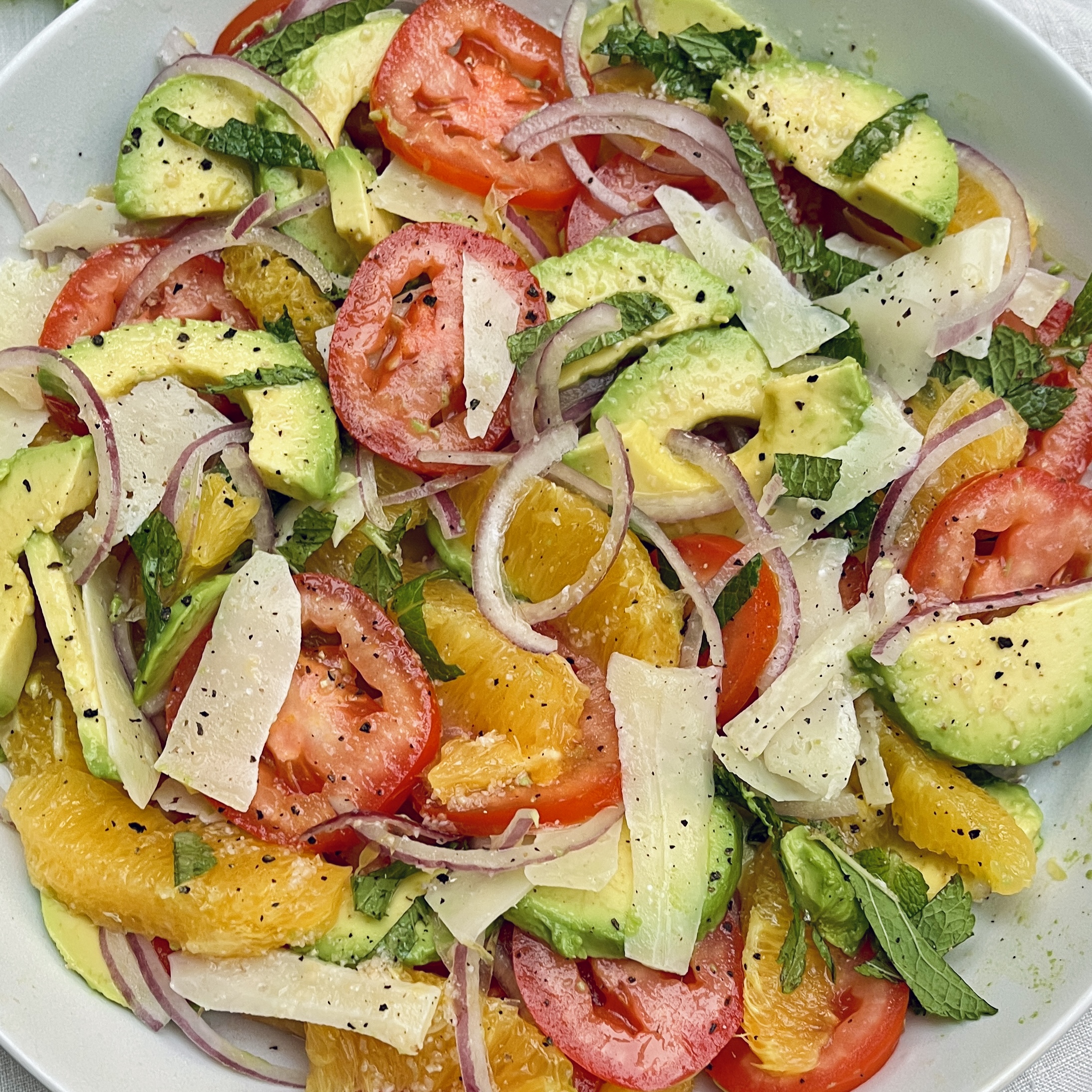 A close up plate of orange, tomato and avocado salad with sliced red onion, chopped mint and shaved manchego