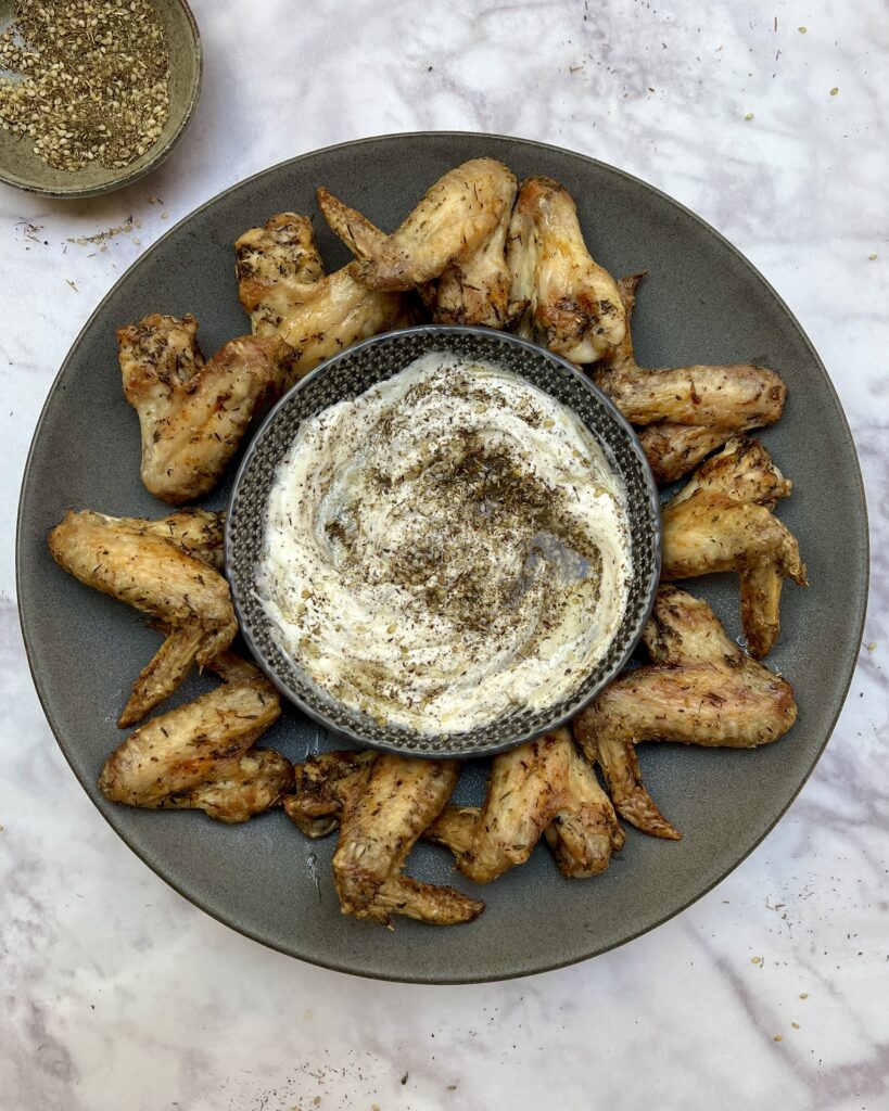 Crispy golden chicken wings arranged on a plate with a bowl with yoghurt and za'atar dressing in the middle