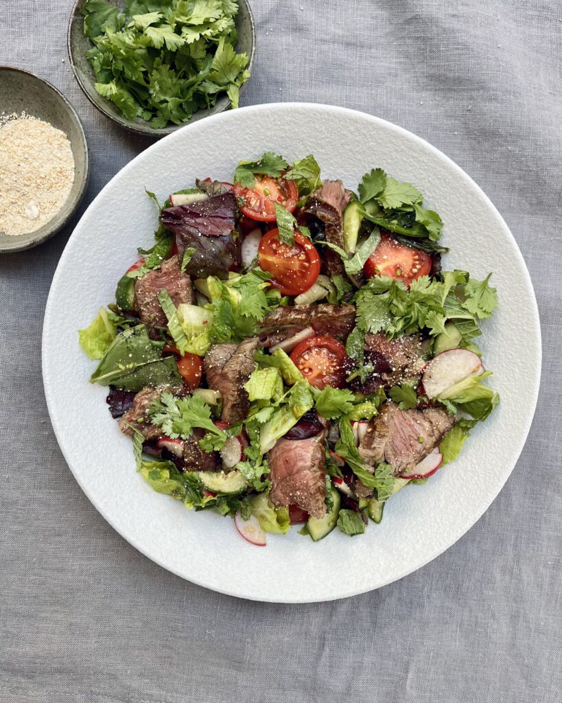 A plate of Thai Beef salad with sliced beef steak, mixed greens, halved cherry tomatoes, radishes and khao khua