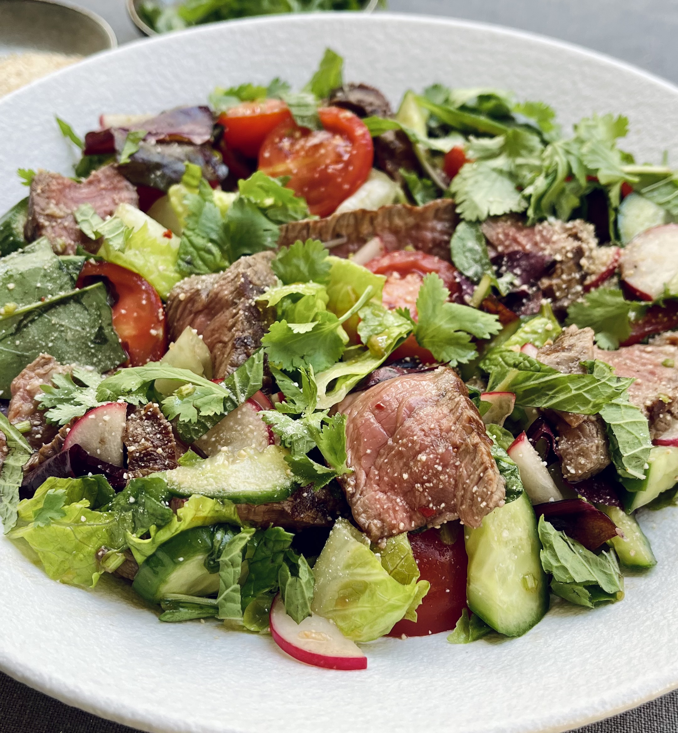 A plate of Thai Beef salad with sliced beef steak, mixed greens, halved cherry tomatoes, radishes and khao khua