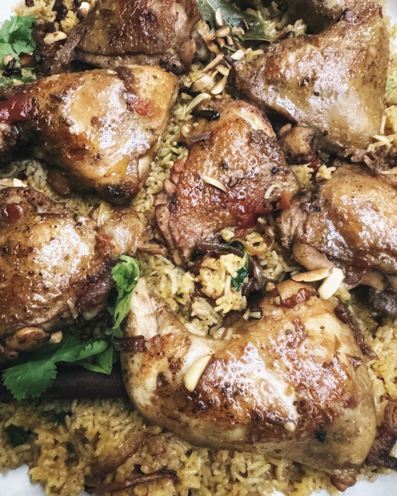 Browned pieces of chicken parts over a bed of dark colored basmati rice garnished with fried onions, coriander leaves and almonds