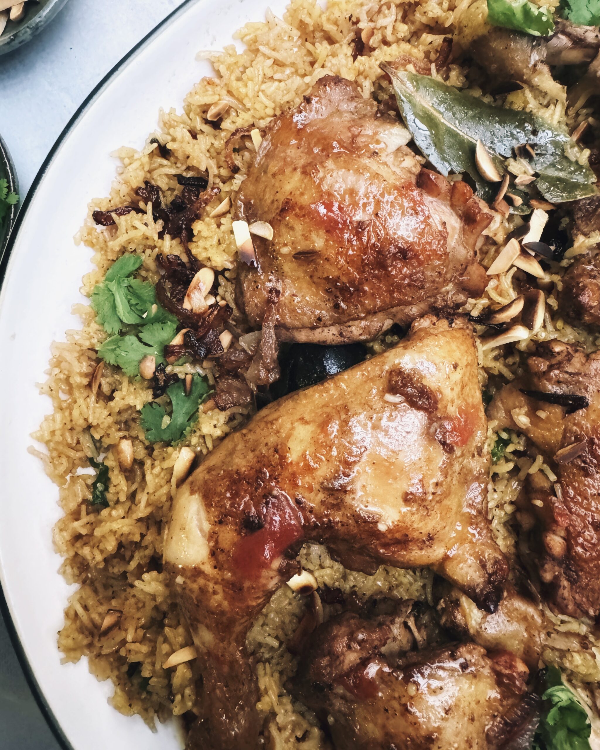 Browned pieces of chicken parts over a bed of dark colored basmati rice garnished with fried onions, coriander leaves and almonds