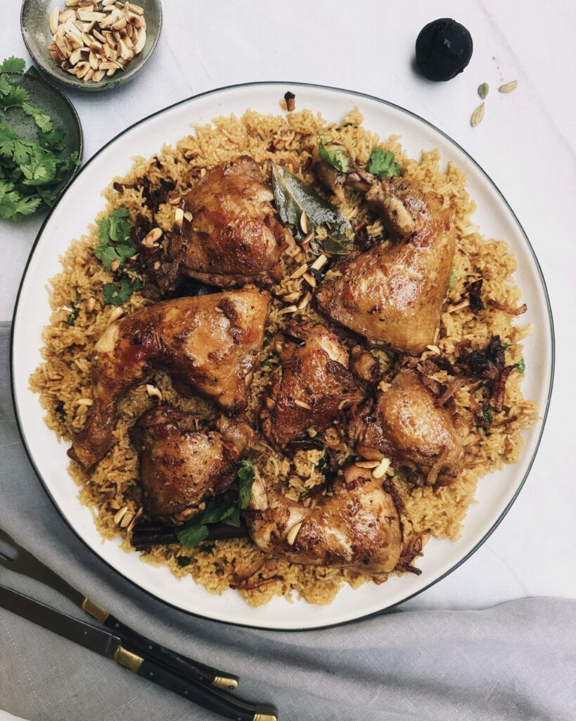 Browned pieces of chicken parts over a bed of dark colored basmati rice garnished with fried onions, coriander leaves and almonds