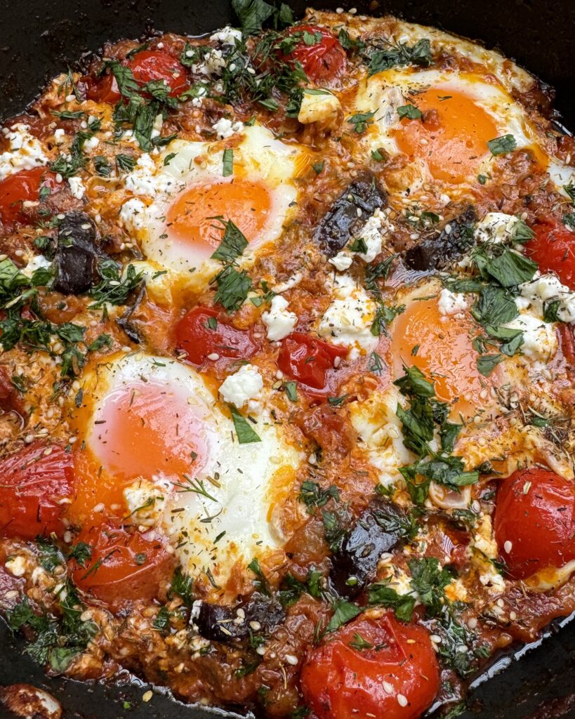 a close up on a skillet with eggplant shakshuka, four poached eggs in a rich sauce topped with feta crumbles, fresh chopped herbs and zaatar