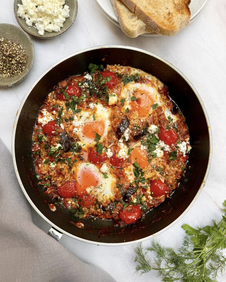 a skillet with eggplant shakshuka, four poached eggs in a rich sauce topped with feta crumbles, fresh chopped herbs and zaatar