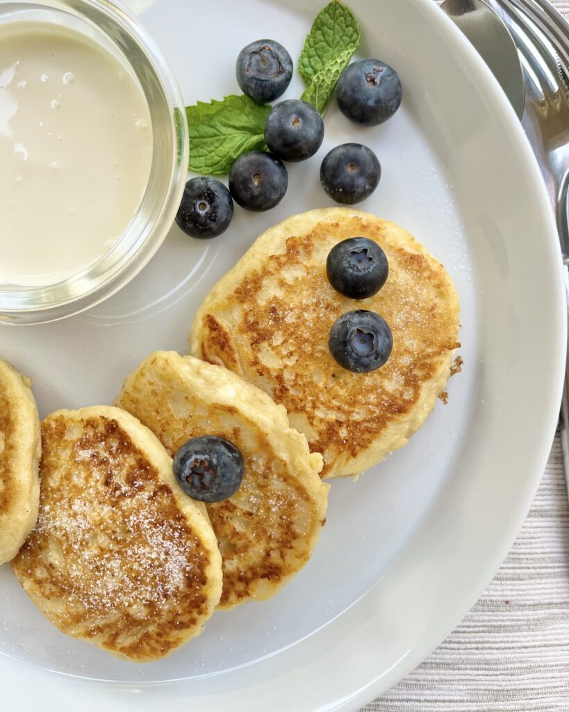 A close up on a plate with syrniki, Russian cottage cheese pancakes, served with sour cream and fresh blueberries