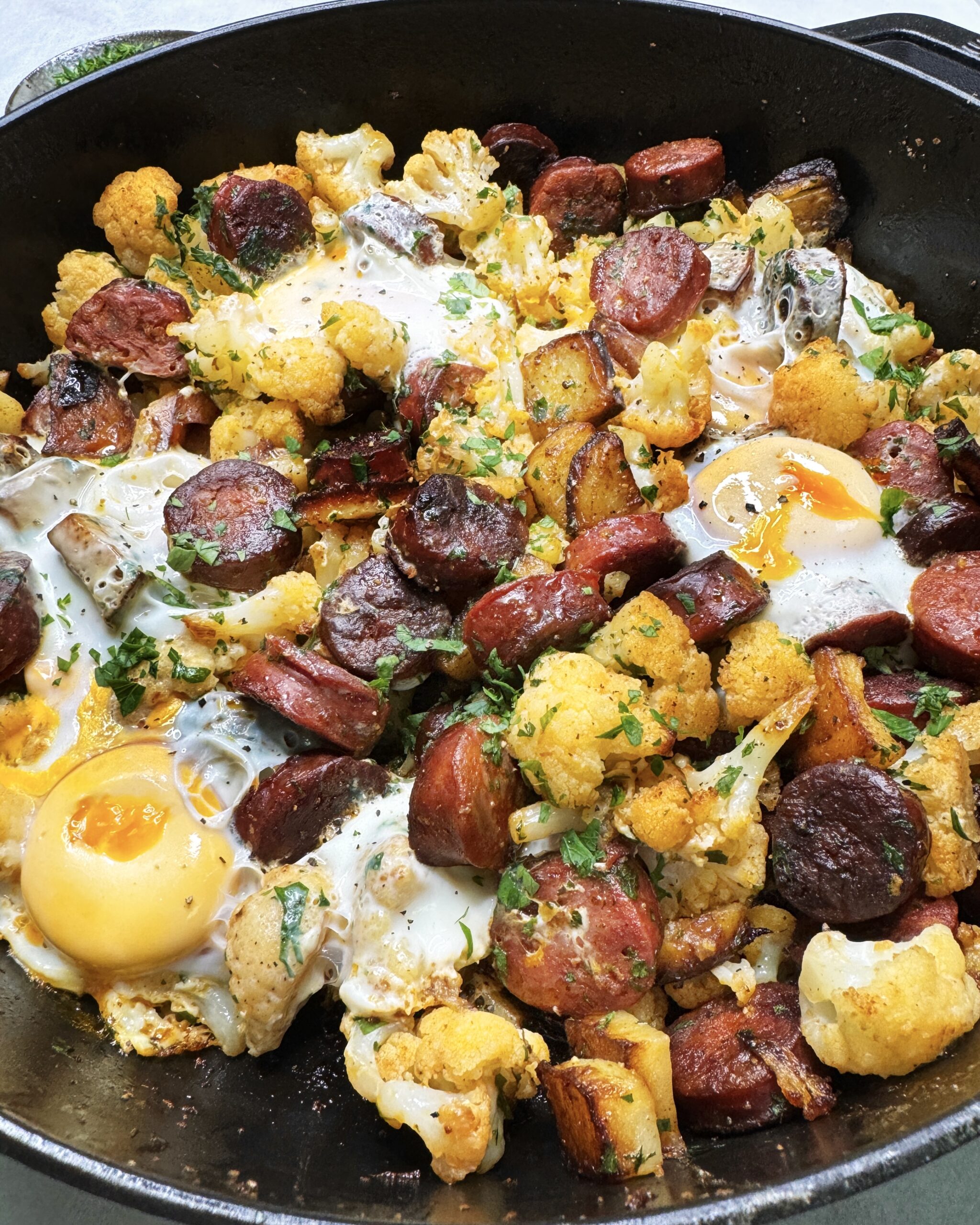 a close up on a skillet with fried cauliflower florets and sliced chorizo topped with fried egg and fresh chopped parsley