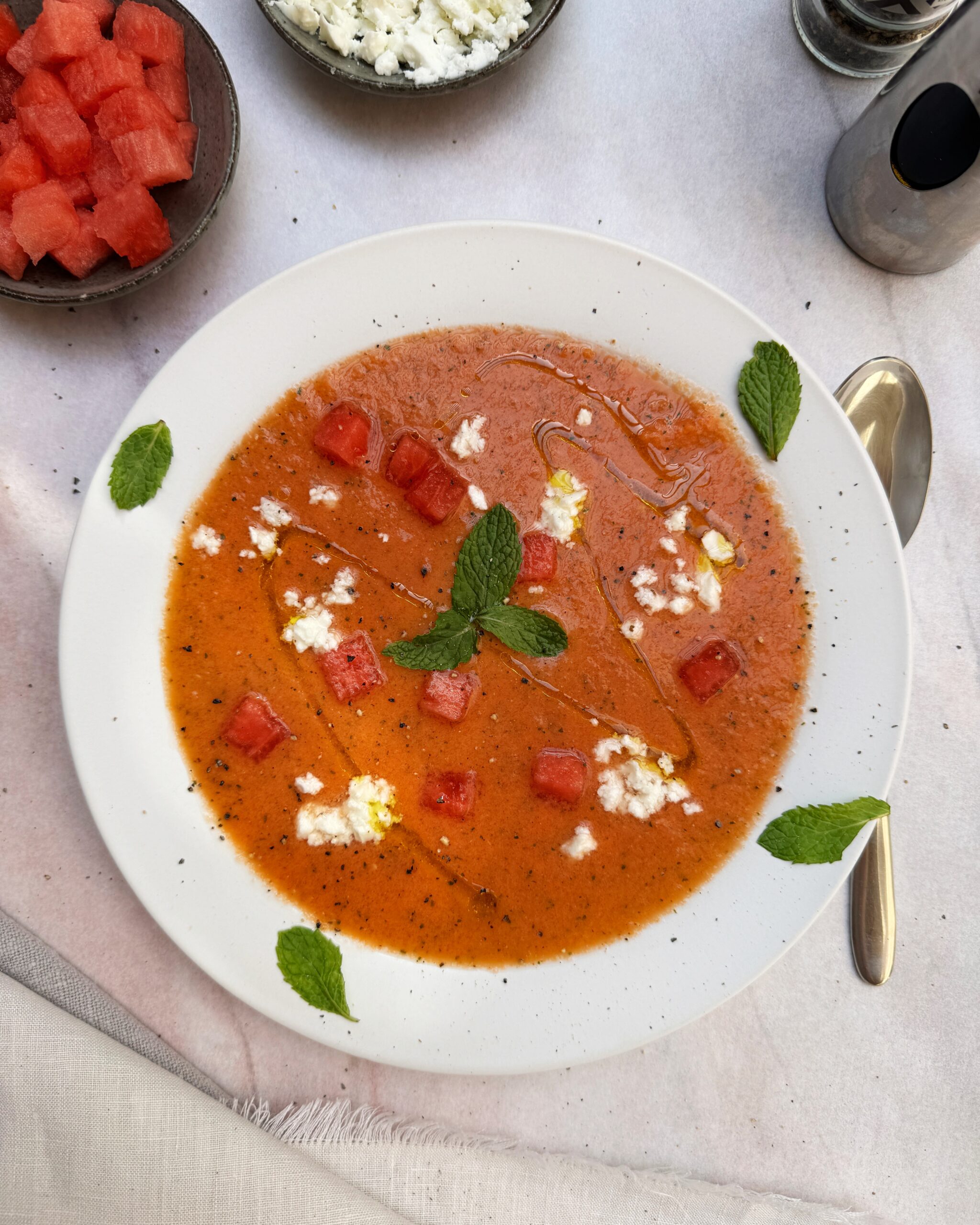 A plate with Watermelon Gazpacho garnished with watermelon cubes, crumbled feta, mint leaves and black pepper