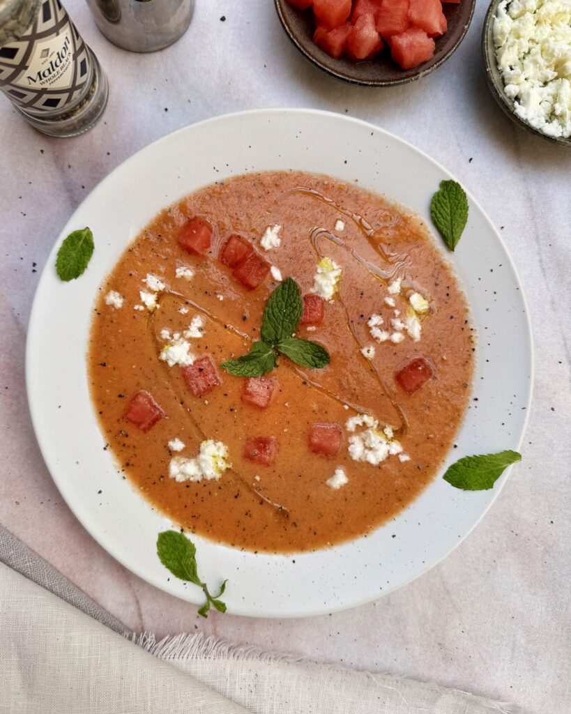 A plate with Watermelon Gazpacho garnished with watermelon cubes, crumbled feta, mint leaves and black pepper