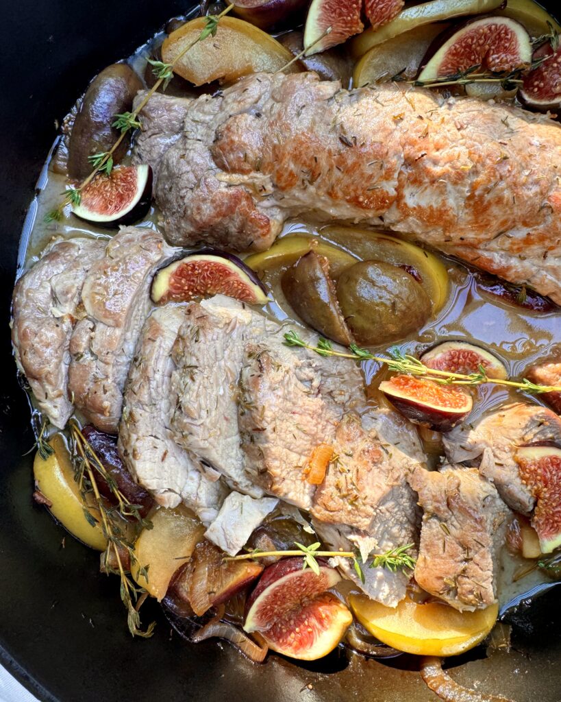 A close up on pork tenderloin in a large black casserole with stewed apples, figs and thyme sprigs