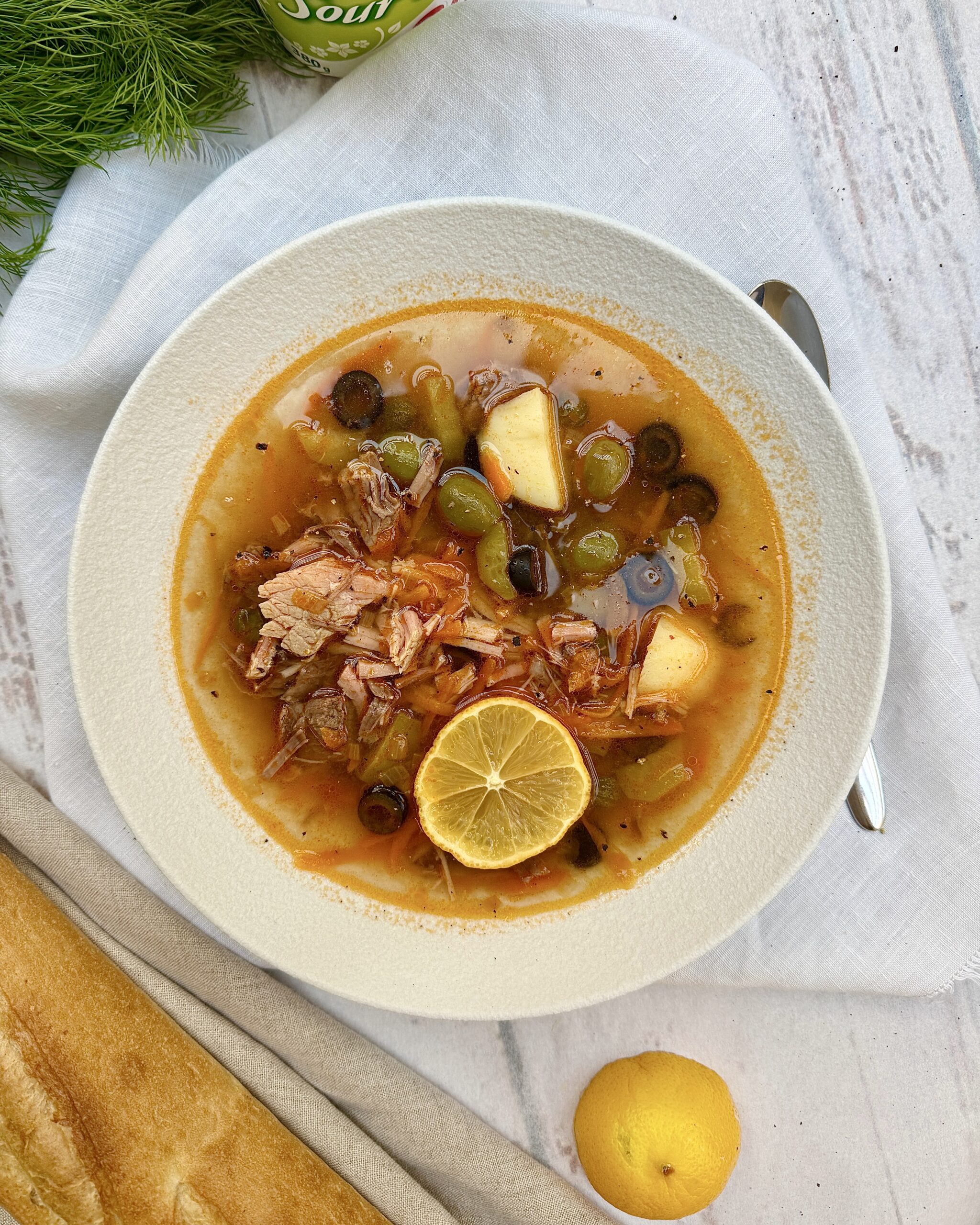 A bowl of meat solyanka soup with pieces of meat, olives, capers, potatoes and lemon